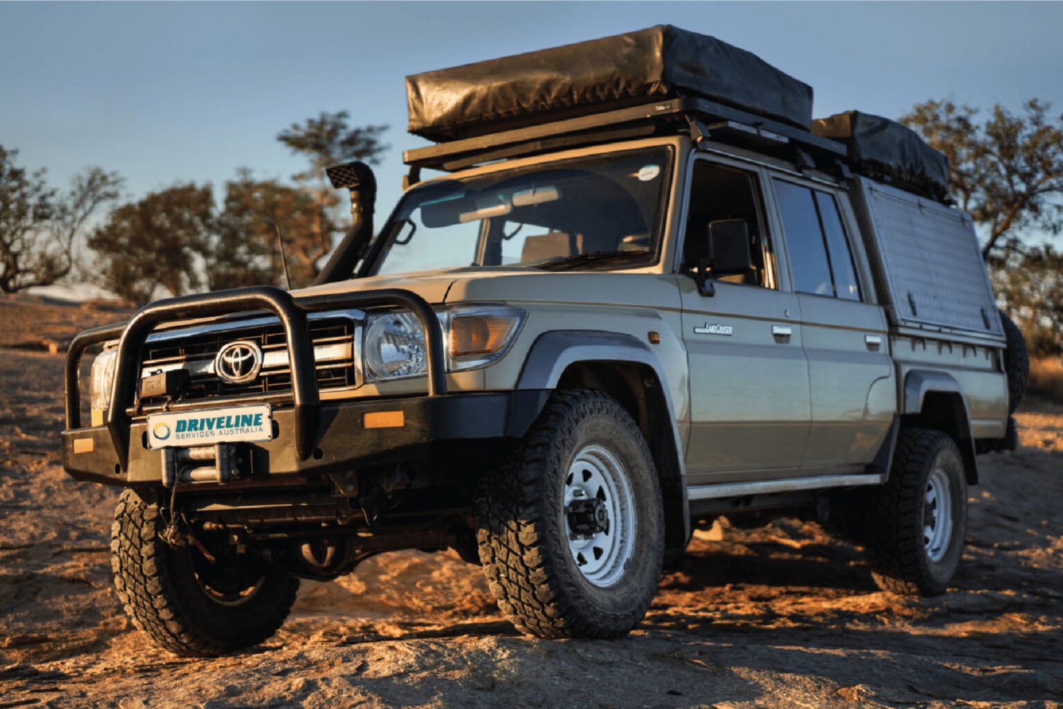 Toyota LandCruiser 70 Series wagon on red dirt outback track at golden hour, fitted with bull bar, snorkel and roof tent